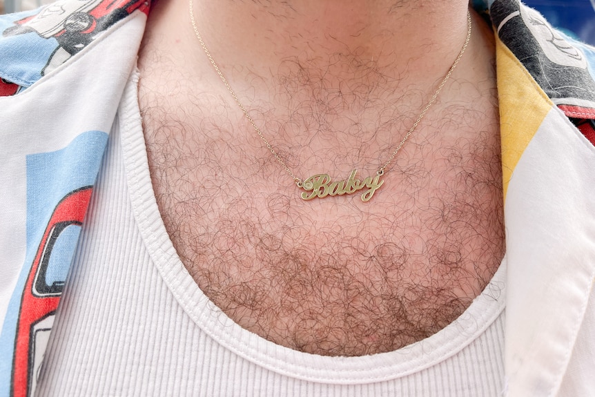 Thomas's gold necklace reads "Baby", seen over a printed shirt over a white singlet with chest hair poking out underneath.
