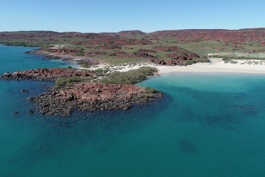 A drone shot of a beautiful beach with blue water, white sand and a collection of red rocks