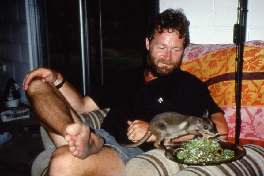 Hogarth sitting on couch with baby wallaby eating grapes off a plate.