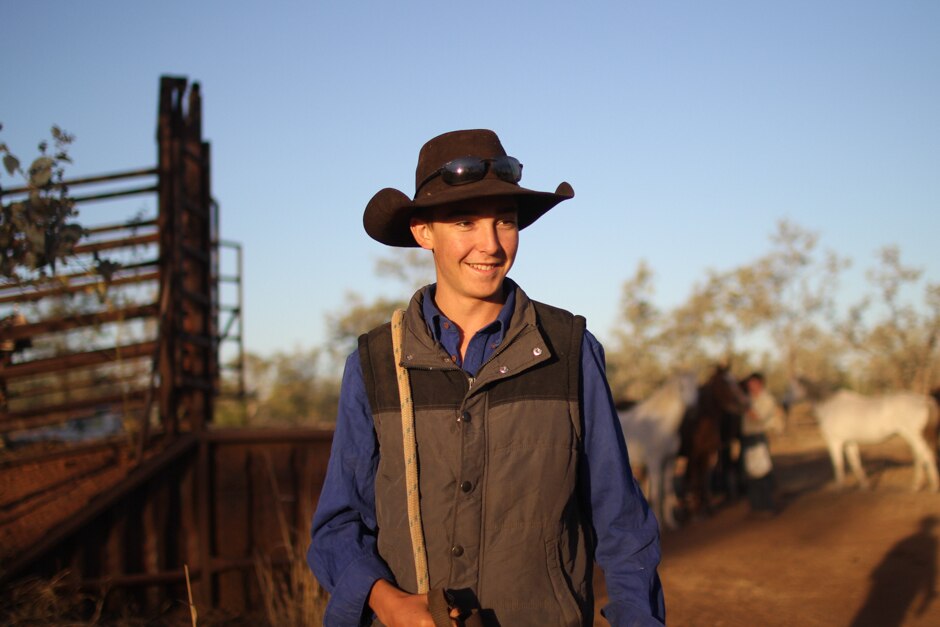 A young man in a wide-brimmed hat with sunglasses on the brim, with horses in the background.