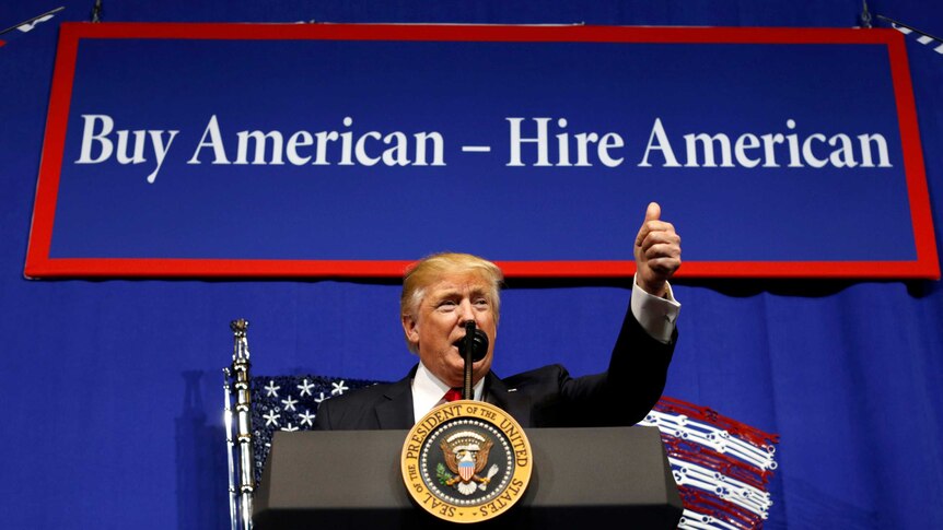 US President Donald Trump stands in front of a buy american hire american sign and gestures with thumbs up