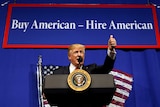 US President Donald Trump stands in front of a buy american hire american sign and gestures with thumbs up