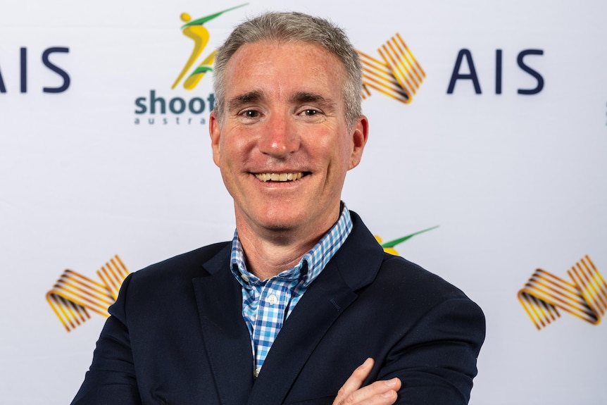 A middle aged man with grey hair in a black suit stands in front of a SA and AIS logo backboard