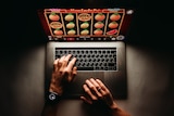 An overhead shot shows a man's hands operating a laptop with a pokie game shown on screen
