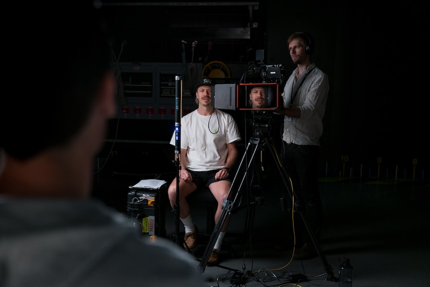 A man standing behind a camera and a man sitting on a chair looking at an interview subject in front of them.