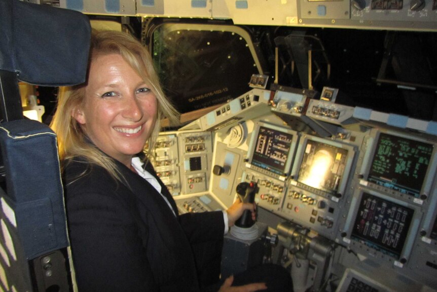 A woman with blonde hair and a wide smile sits in the cockpit of a rocket ship, with her hand on the gear stick.