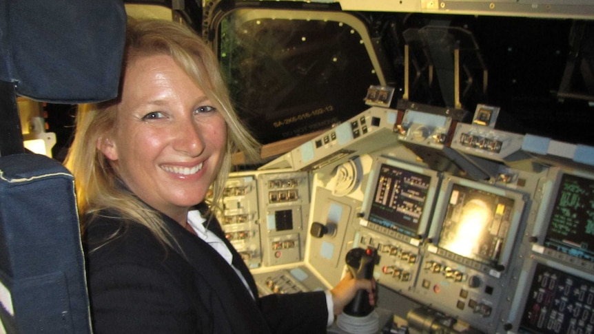 A woman with blonde hair and a wide smile sits in the cockpit of a rocket ship, with her hand on the gear stick.