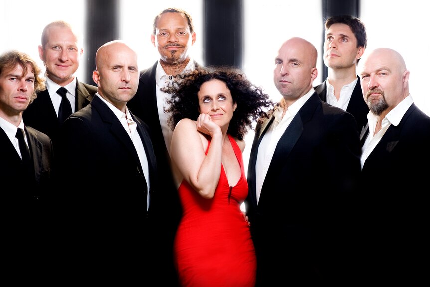 A woman in a red dress surrounded by seven men in suits