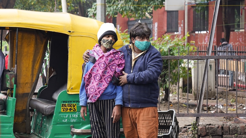 A man holds a boy wrapped in a pink scarf and wearing a mask in front of a taxi.