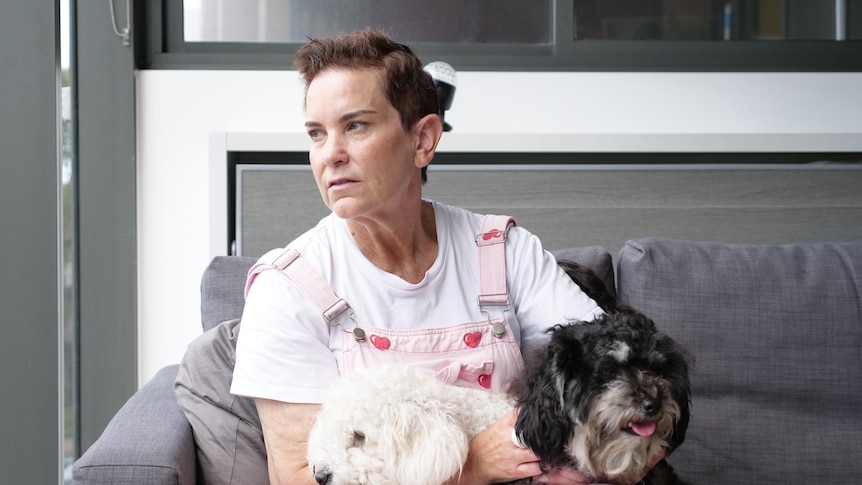 A woman wearing pink overalls over a white t-shirt looks to the right, with two dogs in her lap