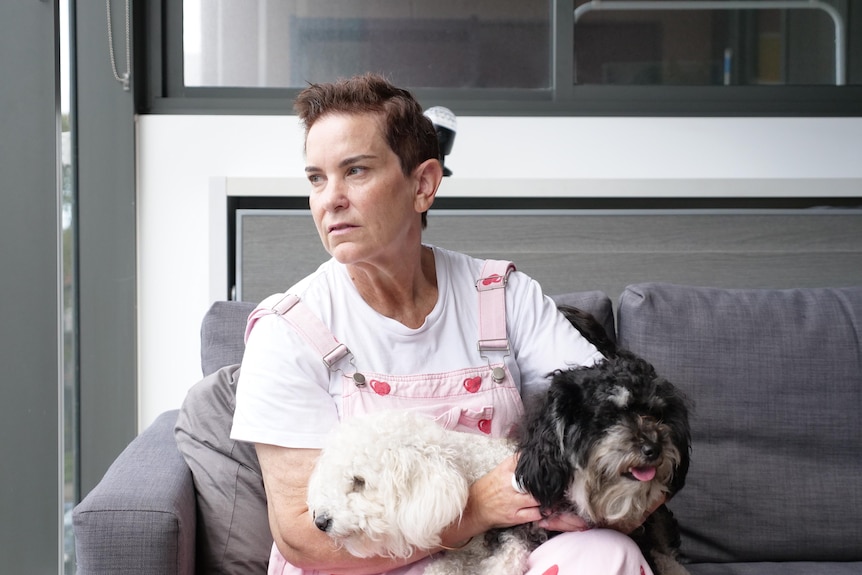 A woman wearing pink overalls over a white t-shirt looks to the right, with two dogs in her lap