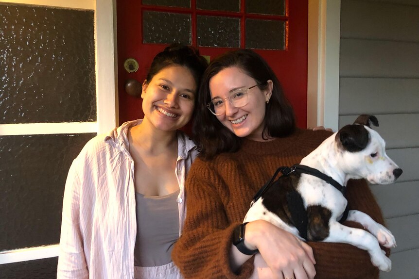 Two young women outside a red front door, smiling and holding a dog. 