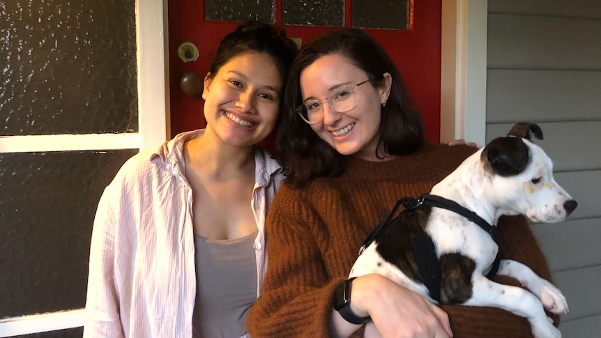 Two young women outside a red front door, smiling and holding a dog, in story about going to friends' places uninvited.