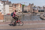 A man in a shirt and jeans rides in bicycle along a bridge during the day in a Medieval-looking city.