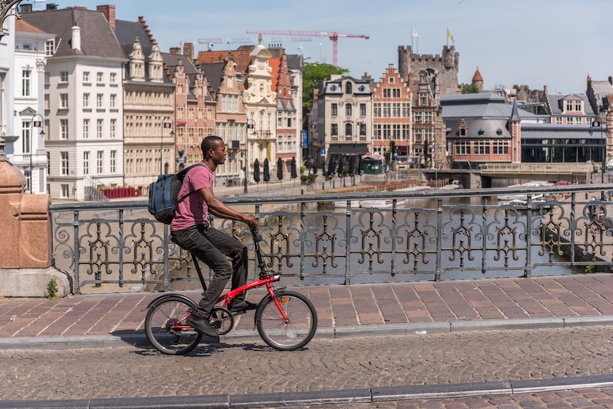 A man in a shirt and jeans rides in bicycle along a bridge during the day in a Medieval-looking city.