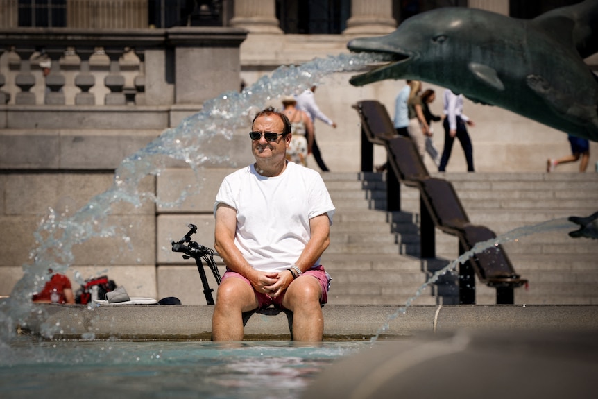 A man in a white t-shirt and shorts sits at the edge of a fountain with a stream of water streaming from a dolphin scultpture