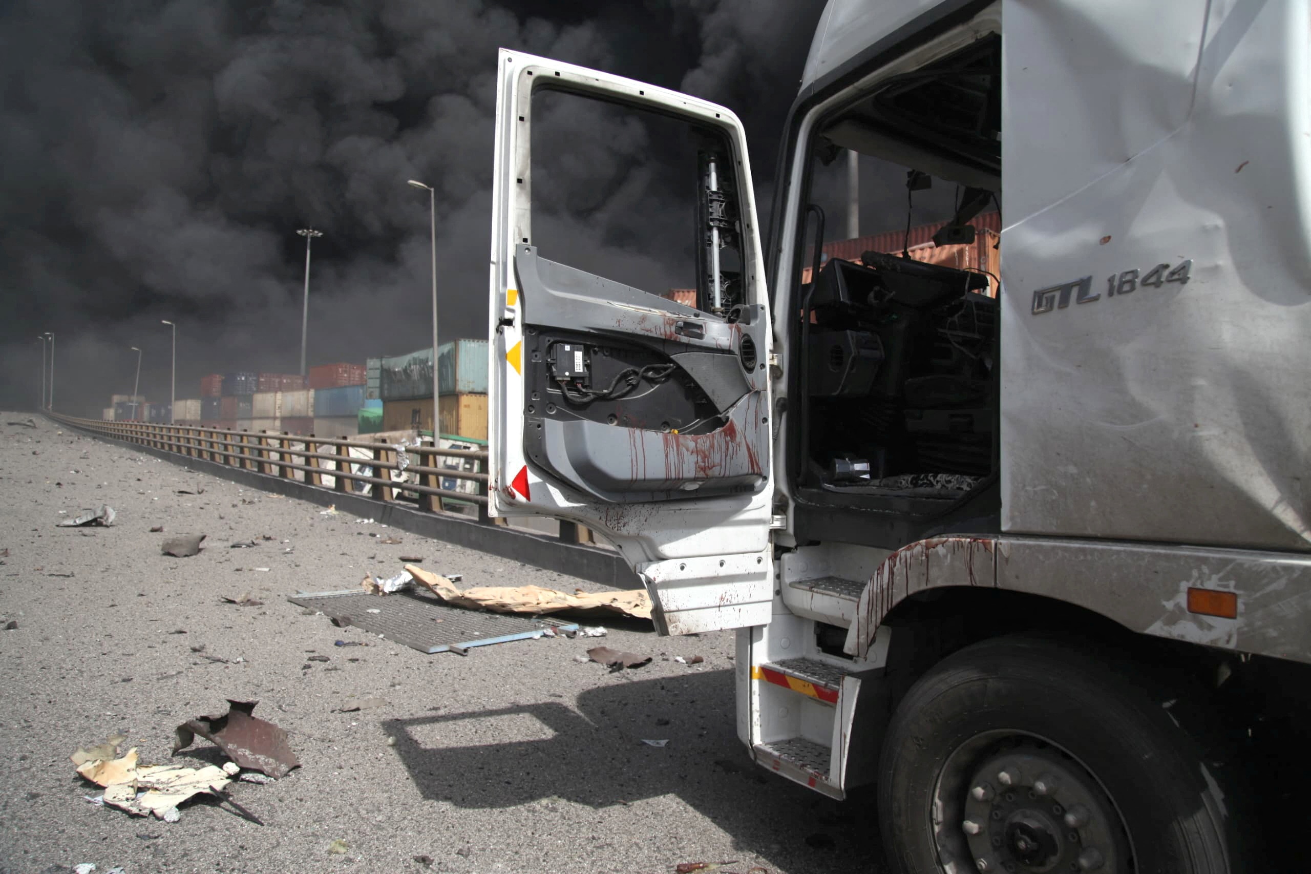 An abandoned truck on a road with smoke that is seen between the shipping containers