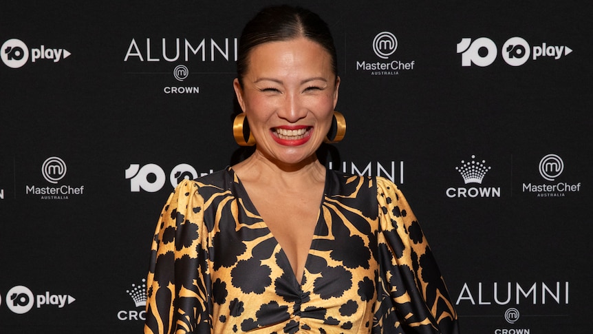 An Asian woman with black hair in a gold and black dress smiles in front of a black background