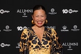 An Asian woman with black hair in a gold and black dress smiles in front of a black background