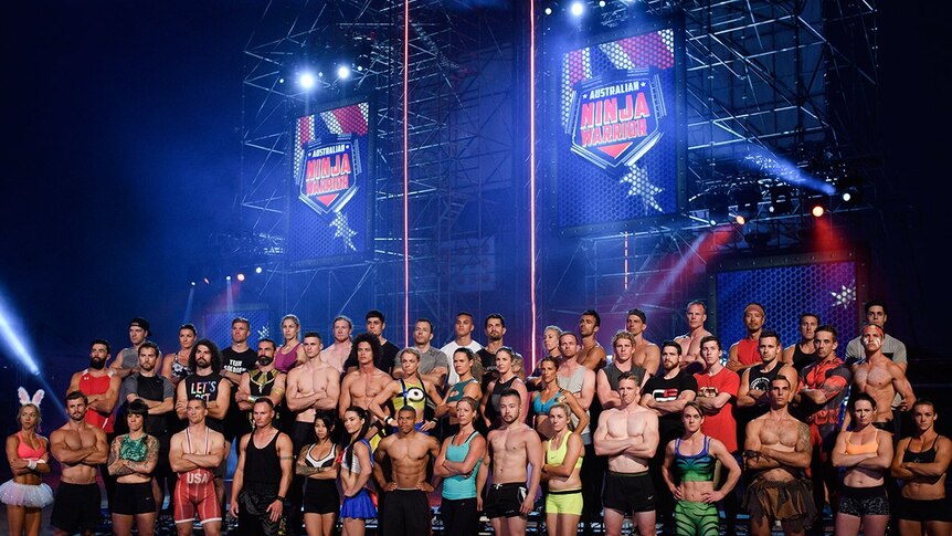 A large group of athletic people pose in front of scaffolding, signs read "Australian Ninja Warrior".;