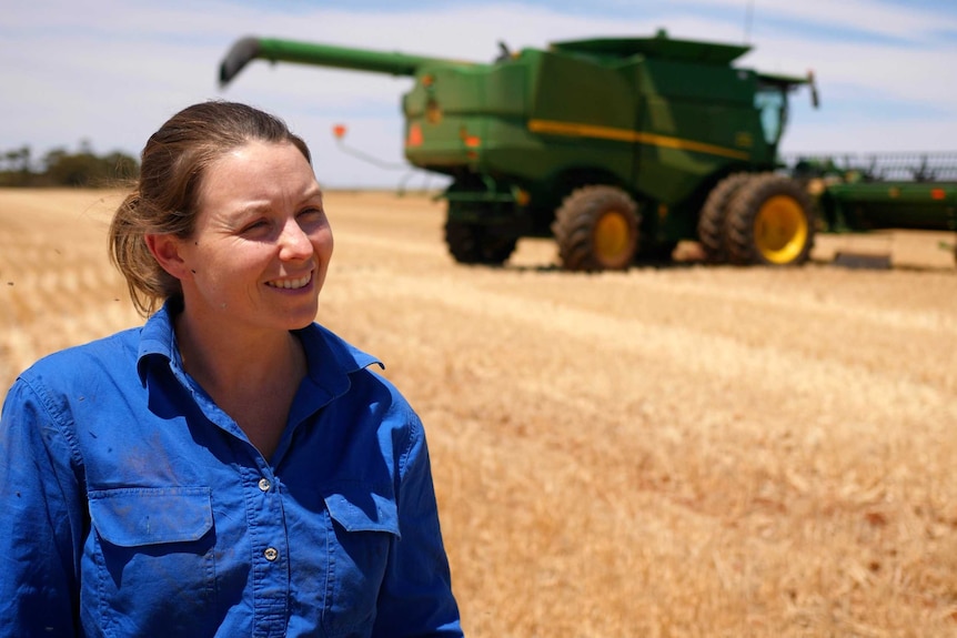 A woman stands in a grain paddock with a green header in the background