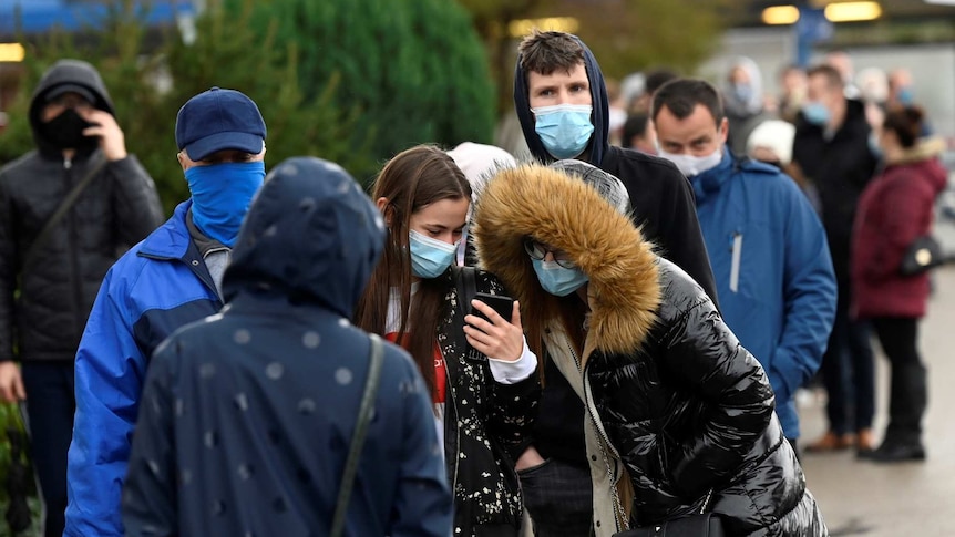 Slovaks wearing masks line up outside as a group infront look a girl's phone