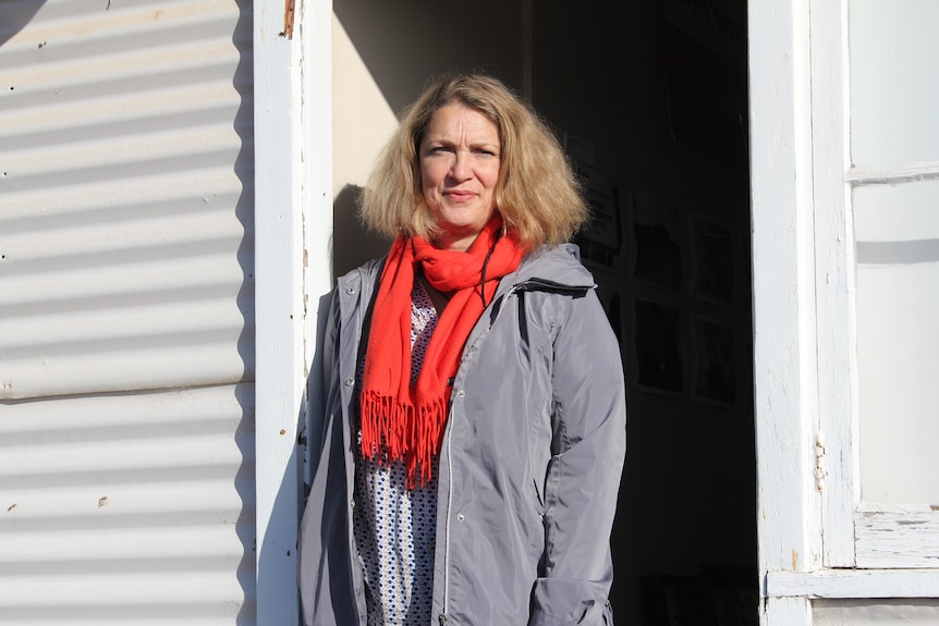 A woman leans against the doorway of a hut clad in corrugated iron.