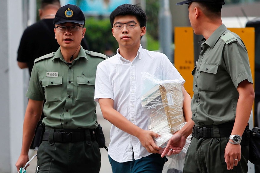 Joshua Wong walks out of prison carrying a stack of books and flanked by two guards.