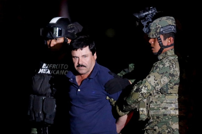 A man in tracksuit pants and a polo shirt is held by soldiers who have bound his hands.