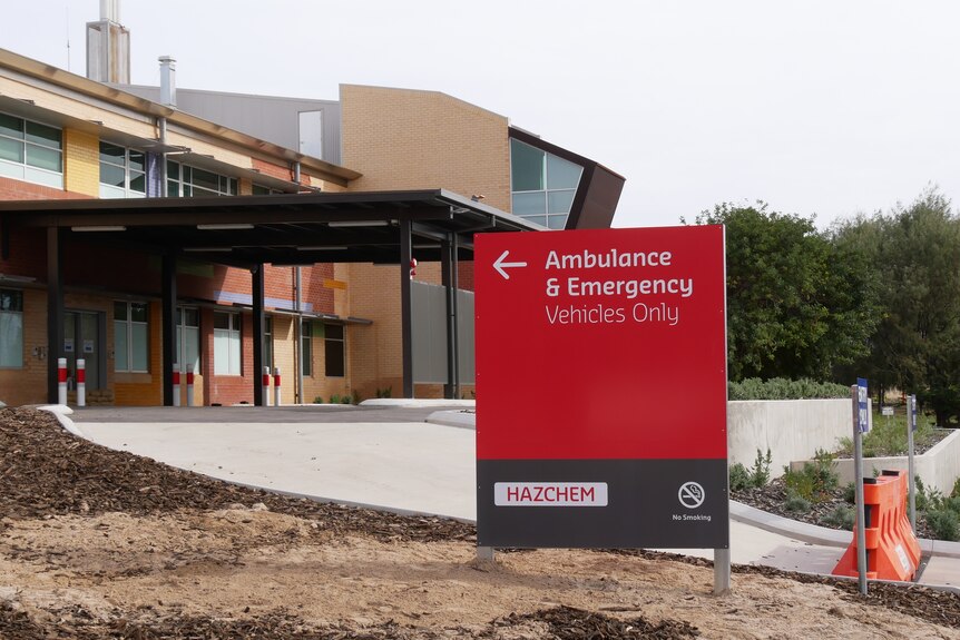 A red sign next to an entrance reads ambulance and emergency vehicles only. Behind is part of a brick hospital. The sky is grey.