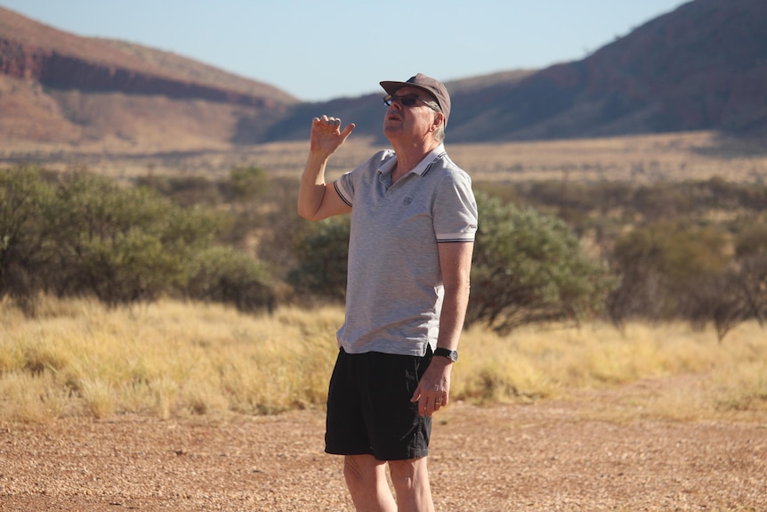 An older man wearing a cap, light t-shirt, dark shorts, stands looking up with the Rawlinson Ranges behind him.
