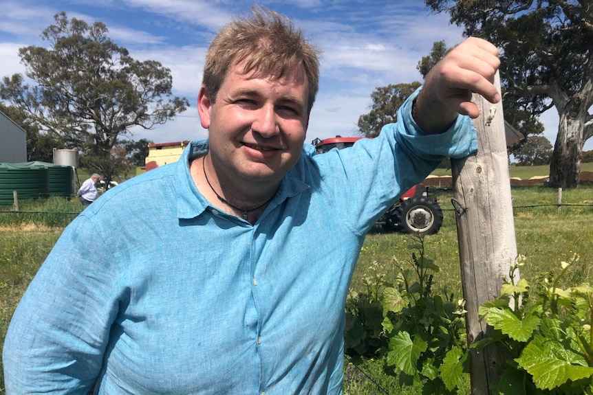 A man in a blue shirt leans against a post and smiles at the camera.