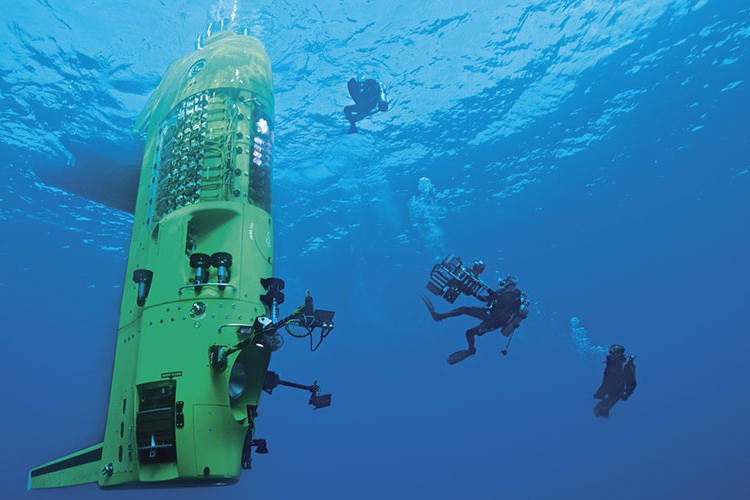 A neon green submersible floating upright in the blue ocean, with divers around it.