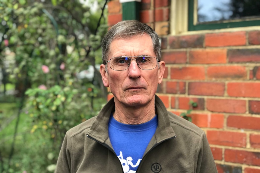James Ashburner, a man with glasses and a blue t-shirt under a khaki jacket, looks seriously at the camera.
