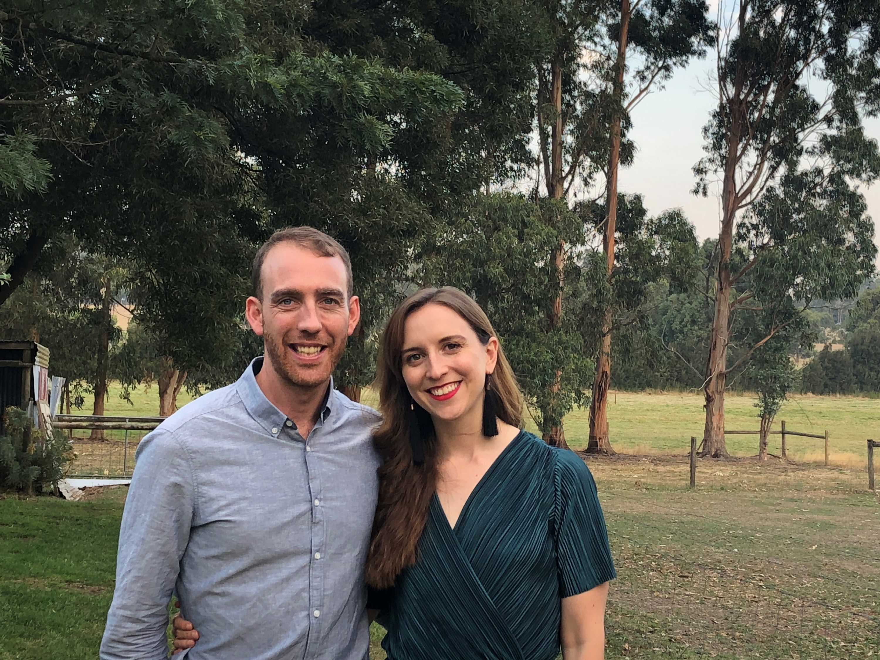 Surrounded by bushland, a man and woman stand close together, arms around one another, smiling.