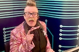 A man in a pink sparkly jacket smiles at the camera with his finger pointed from a home TV studio