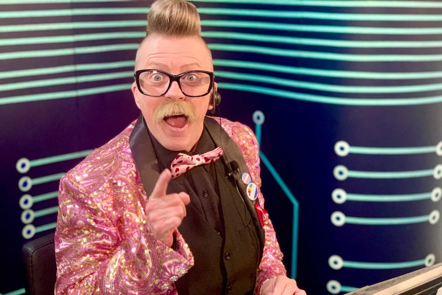 A man in a pink sparkly jacket smiles at the camera with his finger pointed from a home TV studio.