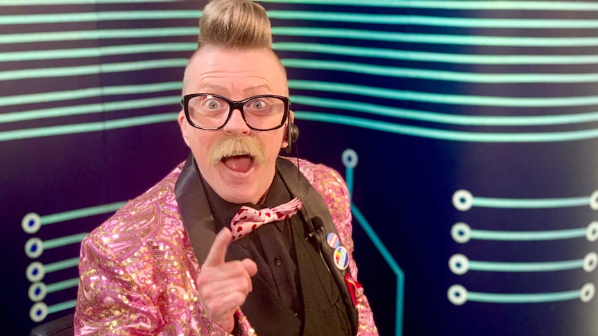 A man in a pink sparkly jacket smiles at the camera with his finger pointed from a home TV studio