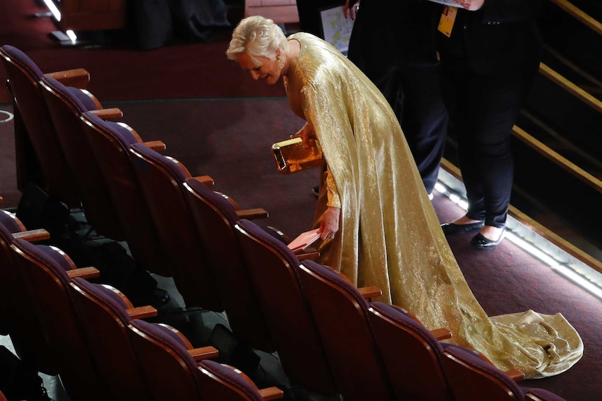 Glenn  Close bends over a theatre seat while wearing a golden gown with a long, flowing cape.