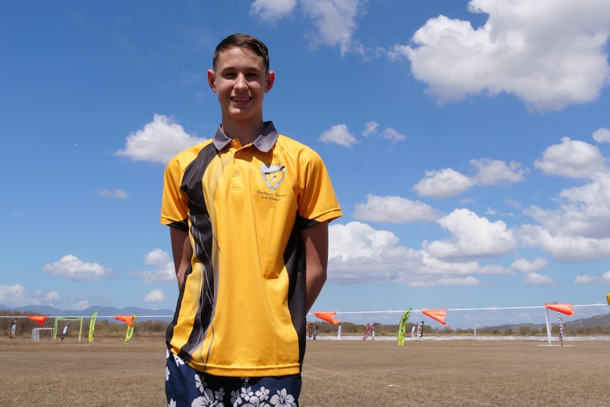 A teenage boy in a yellow Cloncurry State School Shirt stands in front of a field with clouds and sky in the background