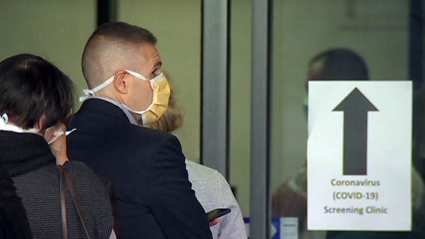 A man wearing a yellow face mask queues up beside a sign for a coronavirus screening clinic.