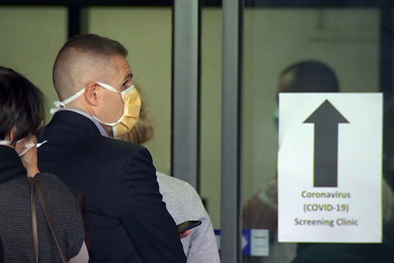 A man wearing a yellow face mask queues up beside a sign for a coronavirus screening clinic.