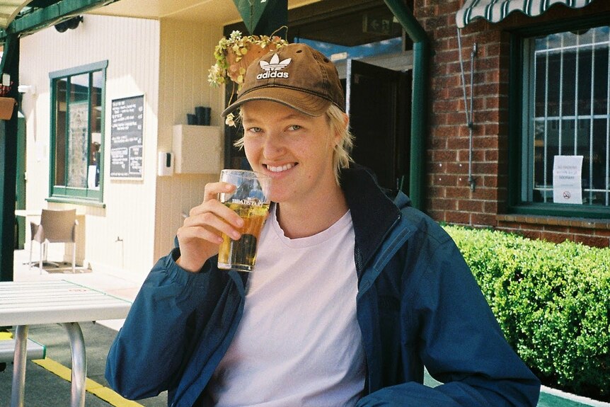A non-binary person holds a schooner of beer and smiles at the camera. They're wearing a cap and white shirt with blue jacket.