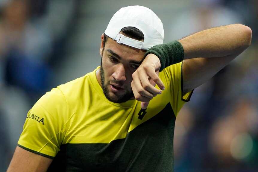 Matteo Berrettini, wearing a yellow and black top and white baseball cap, wipes his brow with a black wrist sweat band