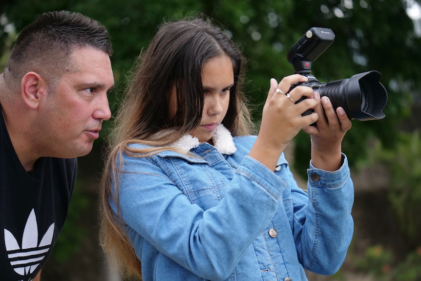 A man leans over a teenage girl's shoulder, while she concentrates on looking into a camera she's holding