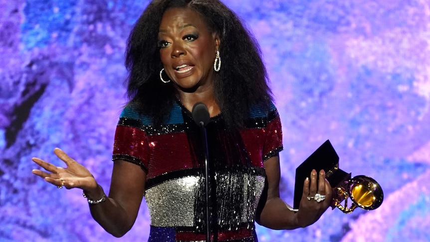 Viola Davis standing, speaking and gesturing with her hands at a microphone, while holding a Grammy award.