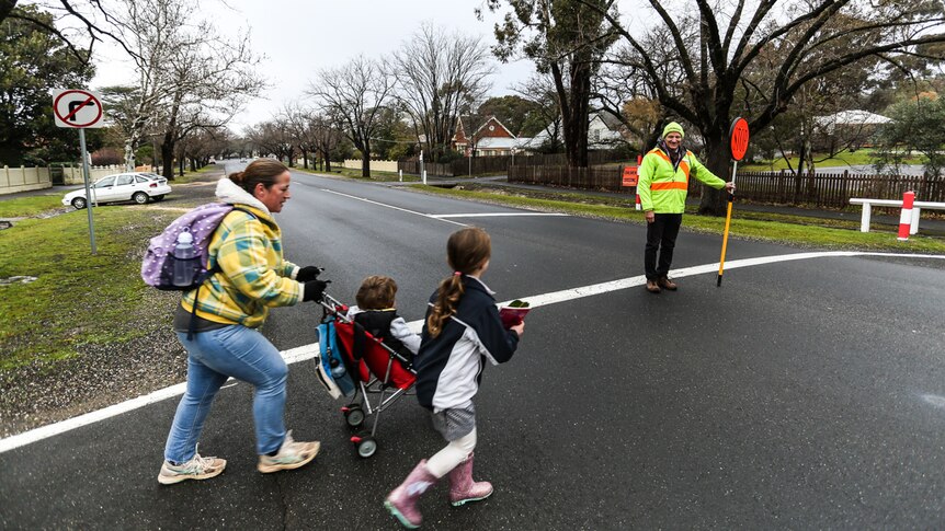 A mother pushes her a pram, and escorts another of her children, across the road. Traffic is halted by a crossing supervisor.