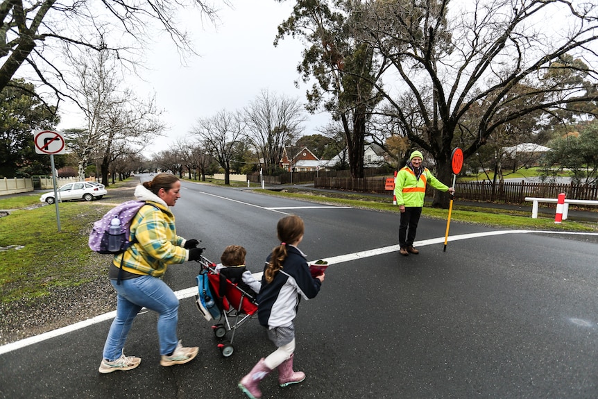 A mother pushes her a pram, and escorts another of her children, across the road. Traffic is halted by a crossing supervisor.