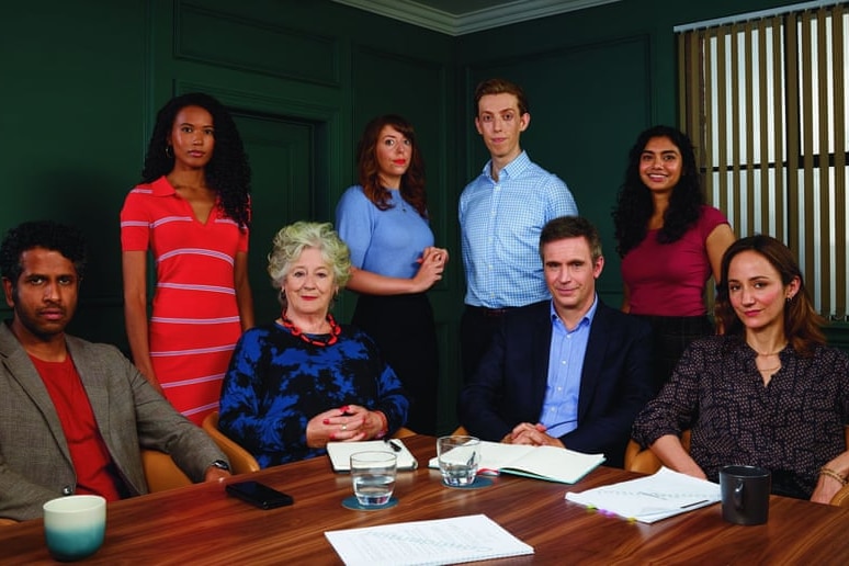 Eight people of various stages sit and stand behind a desk in a conference room. 