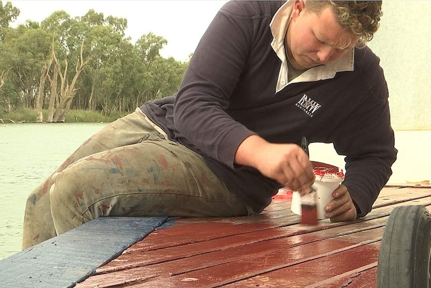 Angus McCullagh painting the deck of the PS Mildura a dark red colour with the Murray River visible behind him.